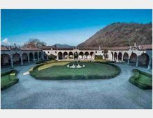 Cortile storico con porticati ad archi su tutti i lati e tetti in coppi, con aiuola ovale di siepi e una piccola fontana centrale, circondato da vialetti in ghiaia. Sullo sfondo si vedono colline boscose sotto un cielo sereno.