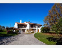 Casa a due piani intonacata di bianco con tetto in coppi rossi e balconi in legno, situata in un ampio giardino. Un vialetto asfaltato conduce all’ingresso, con alberi e vegetazione ai lati sotto un cielo sereno.