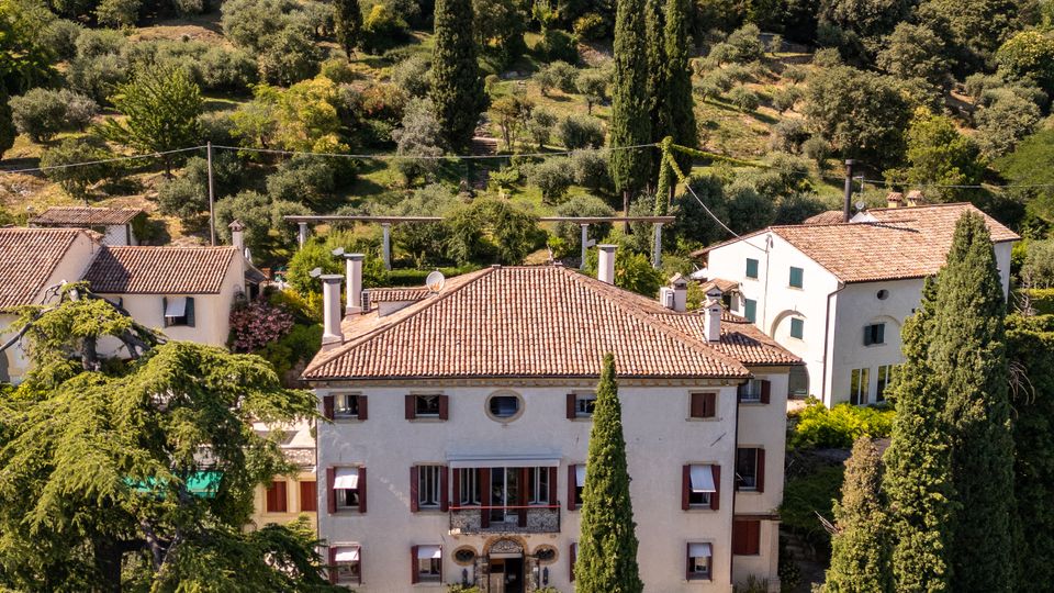Veduta di un borgo collinare con una villa a più piani dai muri chiari e tetto in coppi, circondata da cipressi e altre case. Sullo sfondo, una collina ricoperta di alberi e ulivi culmina in una fortezza in pietra.