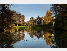 Villa storica in stile neogotico con archi e torrette, situata su un prato oltre un lago. L’acqua calma riflette l’edificio e gli alberi del parco con foliage autunnale.