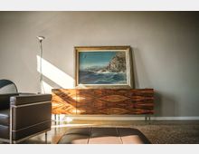 Interno di un soggiorno moderno con parete chiara, credenza bassa in legno venato e grande quadro con paesaggio marino appoggiato sopra. A sinistra si vedono una lampada da terra e un divano in pelle scura, con luce naturale che entra di lato sul pavimento.