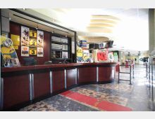 Interno di un cinema con ampia biglietteria in legno scuro e vetri, locandine dei film alle pareti e espositori di snack e bibite sul bancone. Il corridoio di accesso alle sale prosegue sullo sfondo, con pavimento in mosaico e illuminazione a soffitto.