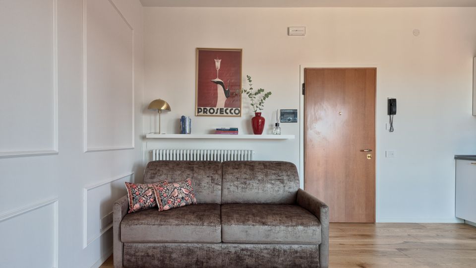 Soggiorno di un appartamento moderno con pavimento in legno, divano grigio e mensola sopra il termosifone con libri, lampada e vaso rosso, sotto un poster “Prosecco”. A destra si vede la porta d’ingresso in legno, il citofono a parete e un angolo di cucina.