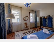 Camera da letto luminosa con due grandi finestre con tende leggere e drappi blu, pavimento in parquet e tappeto decorato. Arredi classici con poltroncina, specchio a parete, lampada da terra, lampadario in vetro blu e armadio aperto con pannelli imbottiti blu.