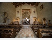 Interno di una chiesa in stile romanico, con navata centrale fiancheggiata da banchi in legno e pavimento in pietra. In fondo si vedono archi in muratura, volte a crociera e l’altare con finestre ad arco e un oculo sopra.