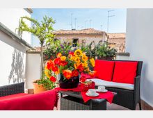 Terrazza su tetto con pavimento in cotto e ringhiera in ferro, arredata con divanetto e poltrona in rattan scuro con cuscini rossi e un tavolino con tazze. In primo piano grandi vasi di fiori colorati e, sullo sfondo, tetti in tegole e antenne sotto un cielo sereno.