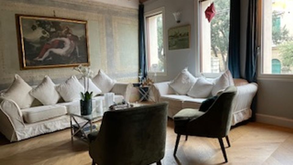 Soggiorno con travi in legno a vista, due divani bianchi con cuscini e tavolini in vetro al centro, affiancati da poltrone scure. Grandi finestre con tende e quadri alle pareti completano l’ambiente.