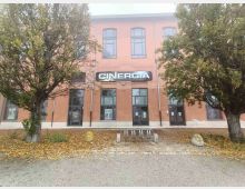 Facciata di un edificio a due piani color salmone con grandi finestre e ingressi vetrati, sormontata dall’insegna “CINERGIA”. Davanti ci sono alberi ai lati, un’area pedonale con foglie a terra e una rastrelliera per biciclette.