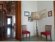 Angolo di un interno domestico con pareti bianche, due sedie in legno con seduta rossa e una lampada da terra con paralume in tessuto, accanto a quadri incorniciati. A sinistra una porta in legno aperta conduce a un corridoio con pavimento lucido e un soffitto decorato da un grande affresco.