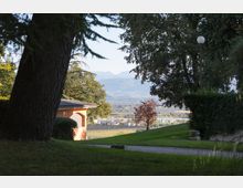 Vista da un giardino in pendio con alberi alti, siepi e un vialetto ghiaioso, con un lampione a globo e un piccolo edificio con portico ad arco. Sullo sfondo si vede una valle con campi e case, e una catena montuosa in lontananza.