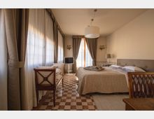 Camera da letto luminosa con grandi finestre a tutta altezza con tende chiare e pesanti drappeggi, pavimento a scacchi e letto matrimoniale con testiera imbottita. Arredi in legno, lampadario centrale, applique a parete e porta-finestra sul fondo con piccola TV su supporto.