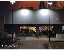 Ingresso esterno di un bar al piano terra di un edificio moderno, illuminato di sera da due lampioni, con ampie vetrate che mostrano tavoli e bancone all’interno. Davanti c’è un vialetto in pietra e alcune aiuole con vegetazione ai lati.