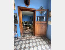 Interno di una casa d’epoca con pareti azzurre, pavimento a piastrelle geometriche e porta in legno con vetri che conduce a un salotto arredato. Sulle pareti sono appesi quadri e una testa imbalsamata, e nella stanza oltre l’ingresso si vedono mobili e decorazioni in stile vintage.