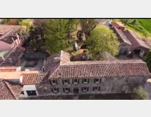 Veduta aerea di un complesso rurale con lunghi edifici in muratura dai tetti in tegole rosse e cortili interni. Intorno si vedono alberi e aree verdi che circondano le costruzioni.