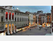 Piazza urbana al crepuscolo circondata da palazzi storici con portici e facciate illuminate, con una colonna monumentale al centro. Sul lato sinistro si vede un edificio neoclassico con grandi arcate e finestre ad arco illuminate di rosso.