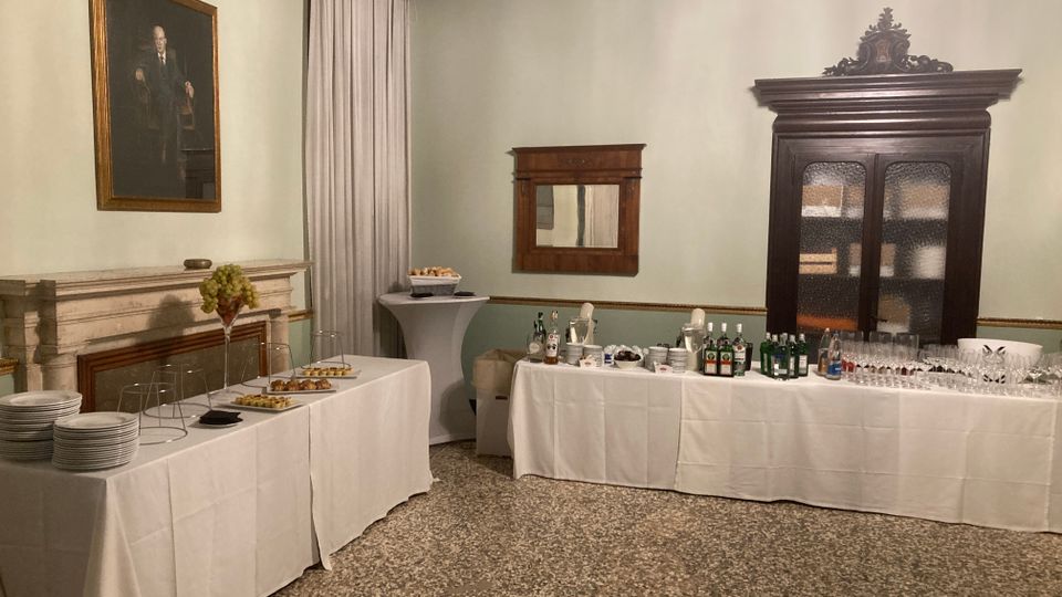 Sala interna in stile classico con pavimento in graniglia, pareti chiare e soffitto decorato, con camino in marmo e ritratto appeso. Lungo le pareti sono allestiti tavoli da buffet con tovaglie bianche, bicchieri e bottiglie, accanto a un grande armadio in legno e una finestra con tende.