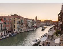 Veduta di un canale urbano con edifici storici dai tetti in coppi e facciate in mattoni e intonaco lungo entrambe le rive, con terrazze e portici sul lungocanale. Diverse imbarcazioni navigano e sono ormeggiate vicino a una riva con tavolini e ombrelloni, mentre sullo sfondo si vedono un campanile e un ponte ad arco.