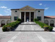 Facciata di una villa storica in stile neoclassico con portico a quattro colonne e frontone, raggiunta da un’ampia scalinata centrale. Ai lati si vedono ali con arcate, tetto in coppi rossi e siepi curate lungo il percorso d’ingresso.