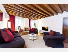 Soggiorno luminoso con soffitto a travi di legno a vista, pareti bianche e pavimento in legno. Divani scuri con cuscini rossi attorno a un tavolino rotondo su tappeto chiaro, con TV su mobile basso e grandi finestre con tende rosse e bianche.