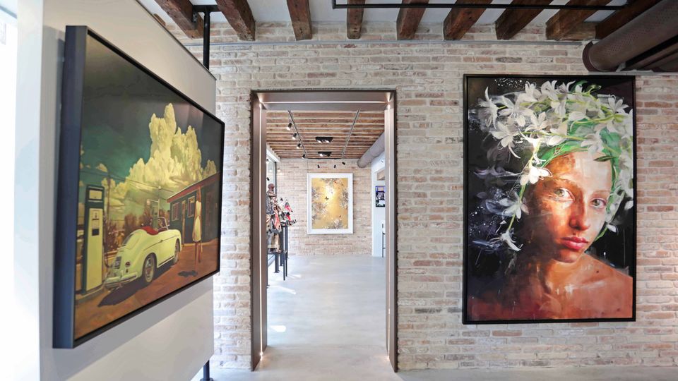 Interno di una galleria d’arte con pareti in mattoni a vista, pavimento in cemento e soffitto con travi di legno e tubazioni industriali. Nel corridoio centrale si vedono grandi quadri appesi alle pareti e un’apertura che conduce a un’altra sala espositiva illuminata da faretti.
