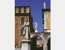 Statue in marmo su alto piedistallo in una piazza storica, affiancata da un arco in pietra decorato e da edifici in muratura con finestre ad arco e persiane. Sul cornicione e sopra l’arco sono visibili altre statue, sullo sfondo di un cielo azzurro.