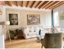Soggiorno luminoso con soffitto a travi in legno, pareti chiare e pavimento in legno. Divano bianco con cuscini, tavolino in vetro, lampada da terra, quadri alle pareti e grandi finestre che illuminano l’ambiente.