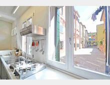 Cucina moderna e luminosa con piano cottura e lavello in acciaio, cappa e utensili appesi, affacciata su grandi finestre. All’esterno si vede una stretta strada pedonale con pavimentazione in pietra e palazzi colorati.