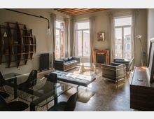 Ampio soggiorno moderno e luminoso con grandi finestre a tutta altezza e soffitto con travi in legno a vista. Arredi contemporanei includono divani e poltrone in pelle scura, tavolo da pranzo in vetro, tavolino in vetro e camino in marmo, con vista su edifici esterni attraverso le finestre.