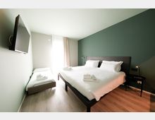 Camera da letto moderna con letto matrimoniale e letto singolo, biancheria bianca e asciugamani piegati. Parete verde scuro con testiera in legno, TV a parete, comodini con lampade e grande finestra con tende che illumina un pavimento in legno chiaro.