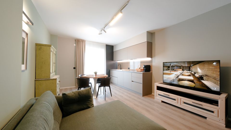 Soggiorno open space moderno con divano in primo piano, tavolo da pranzo vicino a una grande finestra con tende e cucina lineare con pensili e illuminazione sottopensile. A destra c’è un mobile basso con televisore, pavimento in legno chiaro e illuminazione a binario sul soffitto.