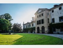Facciata di una villa storica con torre centrale in pietra, finestre con scuri e portico ad archi, affacciata su un ampio prato. Nel giardino si vedono alberi maturi e una piccola fontana circolare vicino al vialetto in ghiaia.
