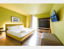 Camera da letto moderna con pareti verde chiaro e pavimento in legno, con letto matrimoniale e comodino. Di fronte ci sono una TV a parete, un mobile contenitore con appendiabiti e una porta-finestra con tende che illumina l’ambiente.