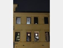 Facciata di un edificio a due piani vista di notte, con finestre illuminate e decorazioni a forma di stella appese all’interno. Sul lato sinistro si intravede una parte di albero decorato con luci dietro una finestra.