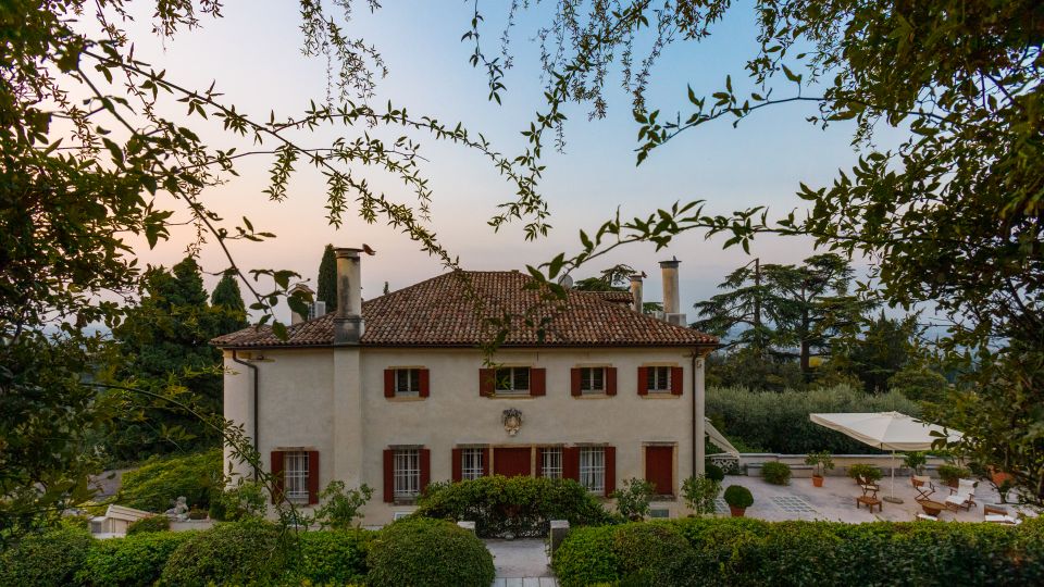 Villa di campagna a due piani con facciata chiara, tetto in coppi e persiane rosse, circondata da giardini curati e siepi. In primo piano si vede un’area esterna pavimentata con ombrellone e sedute, immersa nel verde di alberi e arbusti.