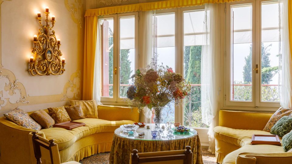 Salotto in stile classico con divani gialli e tavolino rotondo al centro, decorato con vasi e oggetti in vetro. Grandi finestre a tutta altezza con tende leggere e tendaggi gialli si affacciano su un balcone con ringhiera in ferro battuto e alberi all’esterno; pareti con stucchi e applique dorata.