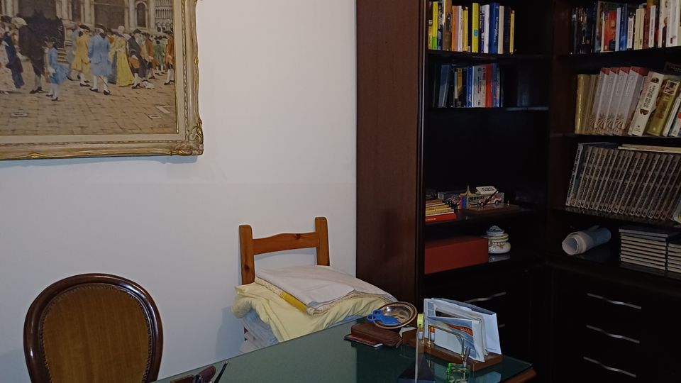 Studio domestico con scrivania in legno e piano in vetro, sedia e blocco appunti, davanti a librerie scure ad angolo piene di libri e raccoglitori. Pareti bianche con quadro incorniciato e lampadario a soffitto, pavimento chiaro con tappeto.