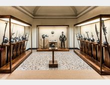 Sala museale con pavimento in seminato e soffitto a volte, con grandi vetrine in legno illuminate che espongono armi antiche e elmi lungo i lati. Al centro c’è una teca con armatura completa e oggetti esposti su piedistalli.