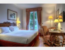 Camera da letto in stile classico con grande letto, testiera in legno intagliato e lampade da comodino, pavimento in legno e scrivania con poltrona a righe. Al centro una portafinestra con tende rosse e bianche affaccia su un giardino verde.