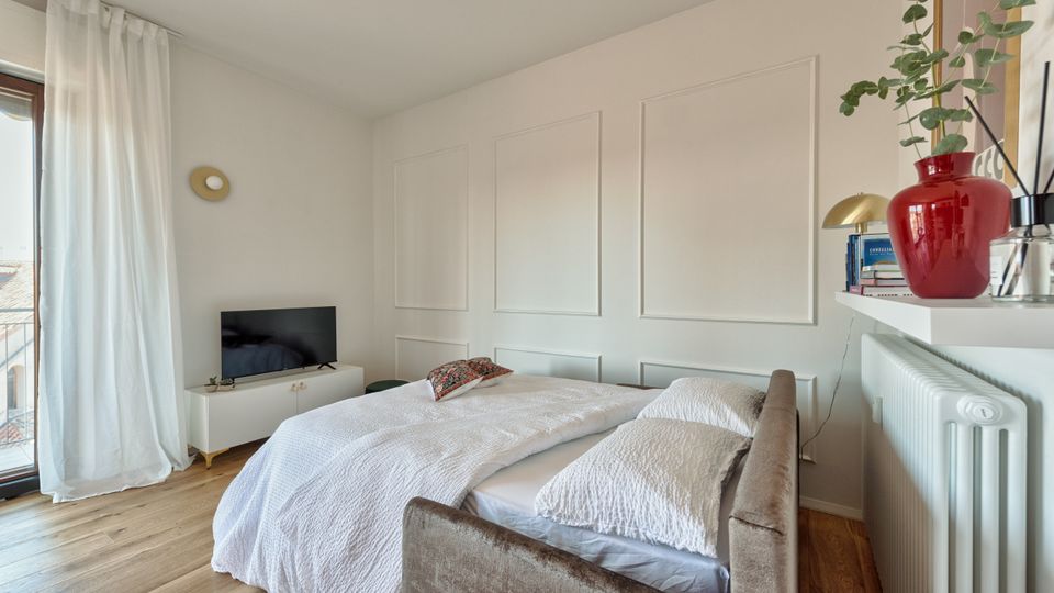 Camera da letto luminosa con pareti bianche a riquadri decorativi, letto matrimoniale con coperta bianca e pavimento in legno. A sinistra una grande finestra con tende leggere e un mobile basso con TV, a destra una mensola sopra il termosifone con libri e un vaso rosso con pianta.
