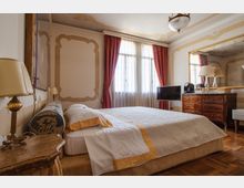 Camera da letto in stile classico con letto matrimoniale, pareti decorate a stucchi e grande finestra con tende rosse e tendaggi bianchi. A lato ci sono un comò in legno con grande specchio, una TV e lampade, su pavimento in parquet e lampadario a soffitto.