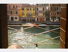 Veduta di un canale urbano con acqua verde e riflessi di luce, osservata attraverso una grata metallica in primo piano. Sulla riva opposta si vedono edifici storici dai colori pastello con persiane e arcate, un piccolo pontile e barche ormeggiate lungo la fondamenta.