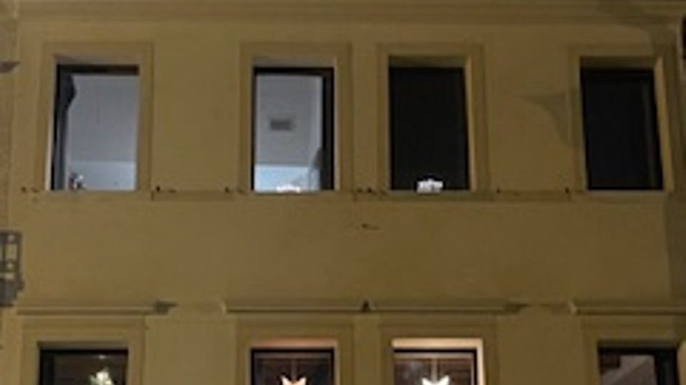 Facciata di un edificio a due piani vista di notte, con finestre illuminate e decorazioni a forma di stella appese all’interno. Sul lato sinistro si intravede una parte di albero decorato con luci dietro una finestra.