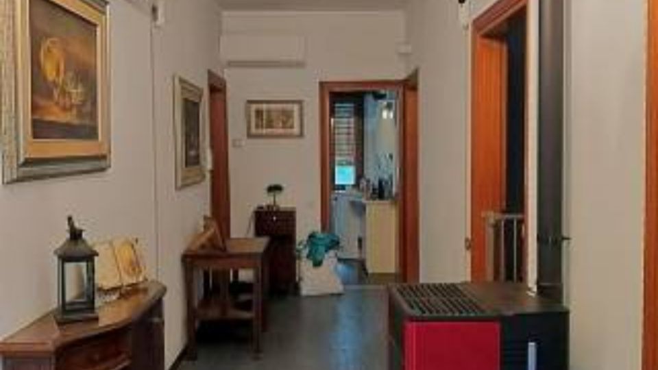 Corridoio interno di un’abitazione con pareti bianche, pavimento scuro e porte in legno che conducono ad altre stanze. Sulla sinistra ci sono quadri e mobili in legno, mentre sulla destra è visibile una stufa a pellet nera e rossa vicino all’ingresso di una stanza.