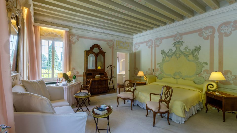 Camera da letto in stile classico con soffitto a travi dipinte, pareti affrescate e grande letto con testiera decorata. Arredi d’epoca, poltrone e divano chiaro, comodini con lampade e ampia finestra con tende che affaccia sul giardino.