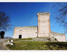 Fortezza medievale in pietra con alta torre quadrata merlata e mura massicce, su un terrapieno erboso. In primo piano si vedono un sentiero, paletti dissuasori e alberi spogli ai lati sotto un cielo sereno.