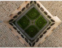 Veduta dall’alto di un chiostro quadrato in muratura con portici ad archi e colonne lungo i lati. Al centro si trova un giardino geometrico con siepi e vialetti di ghiaia, circondato da tetti in tegole.