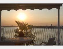 Terrazza coperta con tavolo e sedie in rattan e una palma in vaso, affacciata su un ampio specchio d’acqua con ringhiera metallica. Sullo sfondo si vede la linea dell’orizzonte con edifici in lontananza e il sole basso al tramonto.