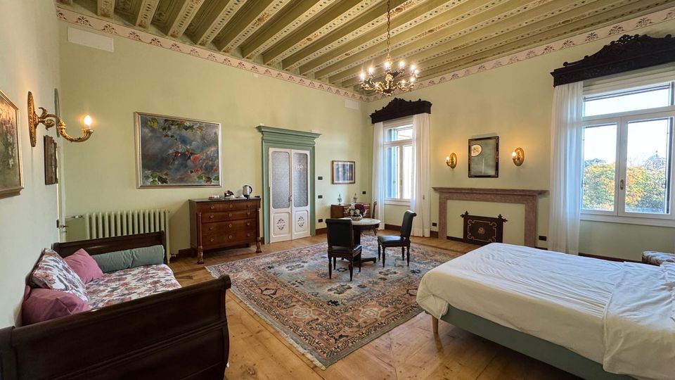 Ampia camera da letto in stile classico con soffitto a travi dipinte, lampadario centrale e pavimento in legno con grande tappeto decorato. Arredi in legno, letto matrimoniale, divanetto, tavolino con sedie, camino e grandi finestre con tende bianche affacciate sull’esterno.