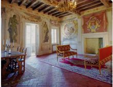 Salone storico con pareti affrescate e decorazioni barocche, soffitto a travi in legno e lampadario centrale. Nella stanza si vedono un grande camino, porte-finestre luminose, pavimento in cotto e arredi d’epoca con tappeti.