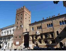 Vista di una piazza con una torre medievale in mattoni con merli e un passaggio ad arco alla base. Accanto si vedono edifici storici con facciate a fasce orizzontali, finestre ad arco e balconi decorati sotto un cielo sereno.